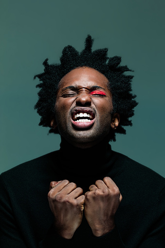 Frustrated afro american man wearing black turtleneck and red make-up on eye, screaming with eyes closed. Headshot on green background.