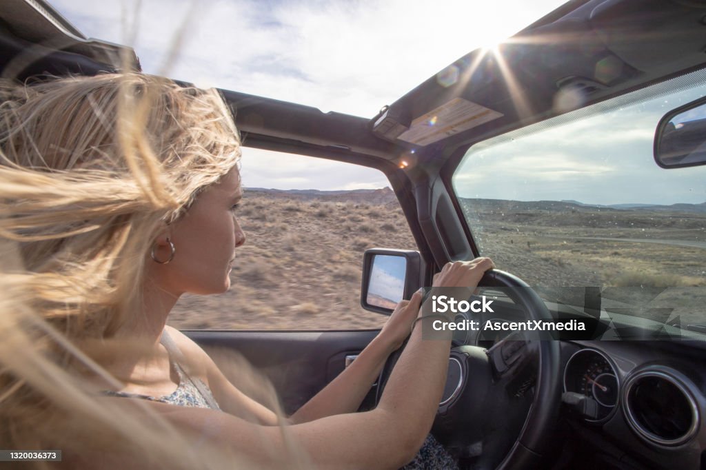 Young woman drives off-road vehicle down desert road She looks out to desert and mountains as wind blows her hair through sun roof Off-Road Vehicle Stock Photo
