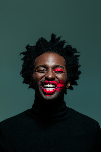 Afro american man wearing black turtleneck and red make-up on eye and lips, laughing with eyes closed. Headshot on green background.