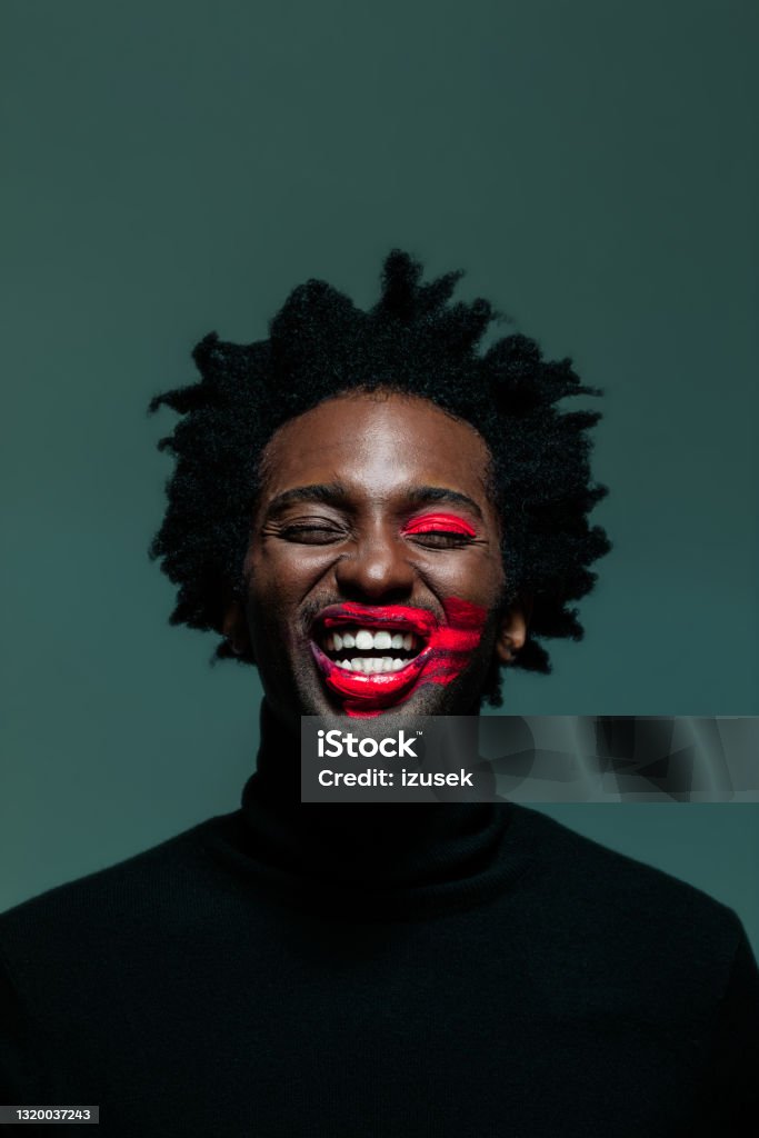 Man with make-up on eye and lips Afro american man wearing black turtleneck and red make-up on eye and lips, laughing with eyes closed. Headshot on green background. Portrait Stock Photo