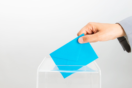 Human hand is inserting blue envelope into ballot box.