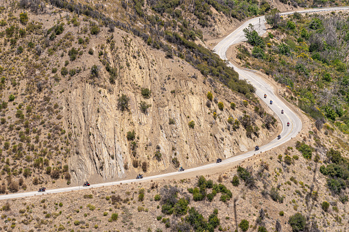 Los Padres National Forest, CA, USA - May 21, 2021: Line of motorcycles ride on road 33 in western part of park in front of beige tall cliff on mountains with green sparse vegetation.
