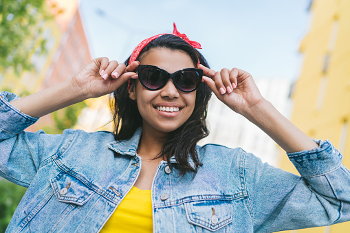 Portrait of cute smiling casually dressed african american student girl posing outdoors in trendy sunglasses and oversized denim jacket.