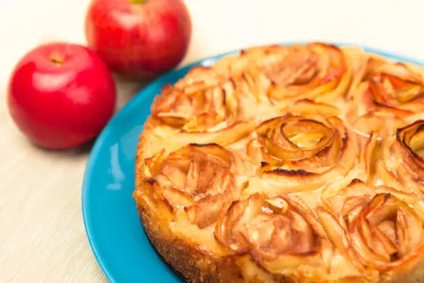 Whole freshly baked apple pie decorated with slices of apple forming roses with two fruits aside.