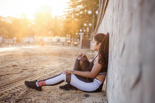 A beautiful black woman sits on the beach. She has just finished a long run and is tired. She is leaning her back up against a concrete wall and one leg is stretched out. She is watching the sunset.