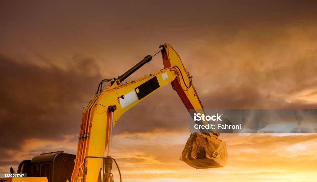 Backhoe working by digging soil at construction site. Closeup hydraulic arm and bucket of backhoe with scoop of soil against yellow sunset sky. Excavator digging on dirt. Trenching machine. Construction Site Stock Photo