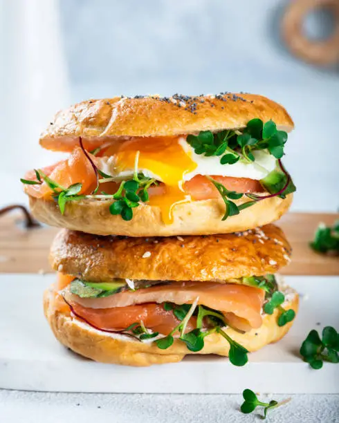 Photo of Healthy freshly baked bagel filled with salmon, microgreen, avocado and egg. Served on white desk. Sandwich with salmon. Healthy breakfast.