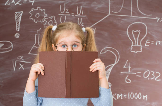 看板と公式でいっぱいの黒板の近くに立って本の後ろに隠れている小さな女の子。幼少期と教育の概念。 - child little girls shy standing ストックフォトと画像