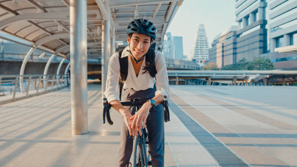 la empresaria asiática va a trabajar a la oficina y sonriente usa mochila mira la cámara con bicicleta en la calle alrededor del edificio en una ciudad. - casco de ciclista fotografías e imágenes de stock