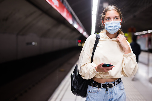 Young beautiful girl in protective face mask standing on the subway platform and using her smartphone