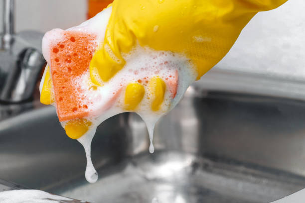рука в желтой латексной защитной перчатке выдавливание пены из оранжевой губки. - protective glove washing up glove cleaning latex стоковые фото и изображения