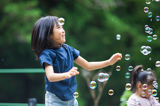 A young Japanese girl is chasing and popping bubbles in the air in her front yard.