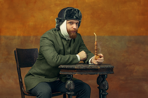 Portrait of red headed and bearded man playing famous artist Van Gogh isolated on dark orange bacground. Concept of art, beauty, eras comparison, fashion. imitation, ad. Bandaged ear.