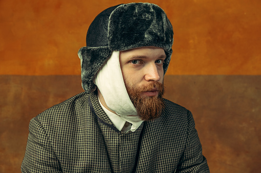 Close-up portrait of red headed and bearded man playing famous artist Van Gogh isolated on dark orange bacground. Concept of art, beauty, eras comparison, fashion. imitation, ad. Bandaged ear.