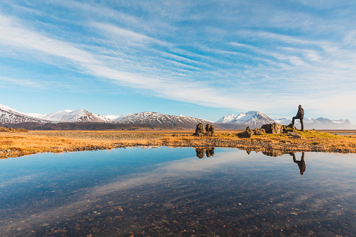 Explorer in Iceland with reflection on the water - Adult man standing on a rock looking around - Majestic mountains on background, reflections on water on foreground - Travel and wanderlust concepts