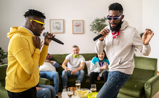 Two friends holding microphone dance and sing while their friends watch from the sofa in a bright modern living room