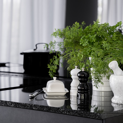 Close-up on black, granite kitchen countertop with decorative, white butter dish, stylish black and white salt and pepper shakers, mortar and fresh, green herbs in flower pots