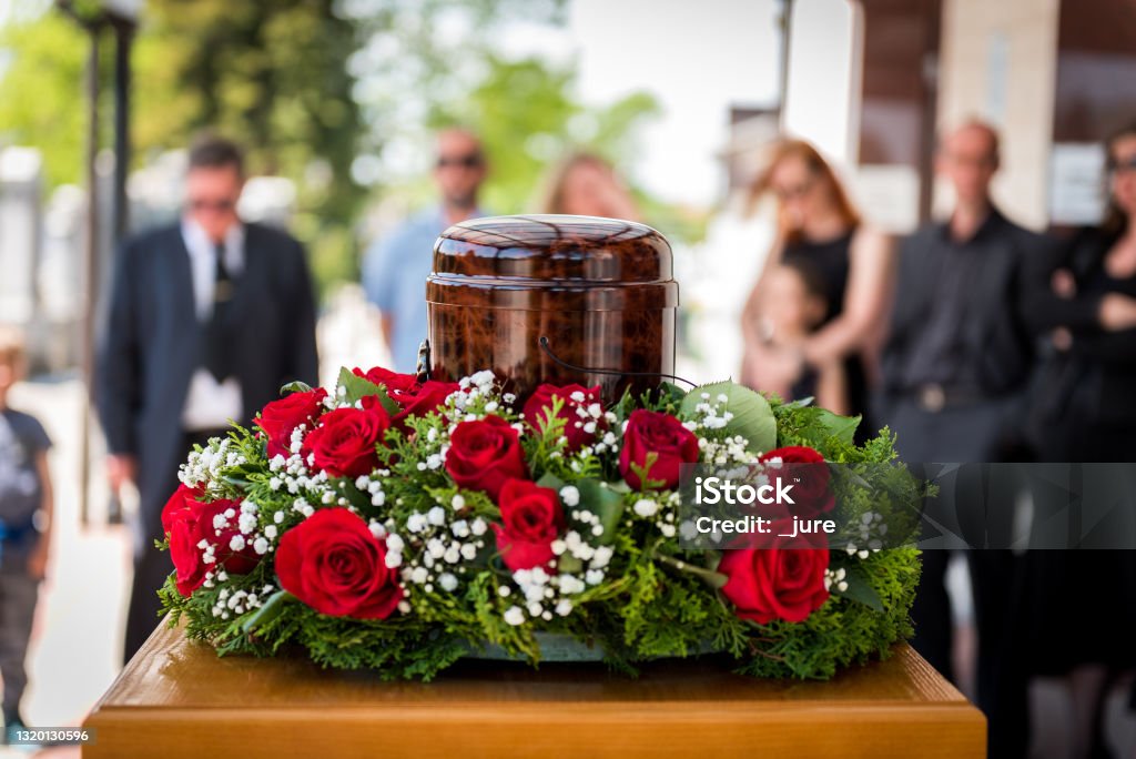Funerary urn with ashes of dead and flowers at funeral. Funerary urn with ashes of dead and flowers at funeral. Burial urn decorated with flowers and people mourning in background at memorial service, sad and grieving last farewell to deceased person. Funeral Stock Photo