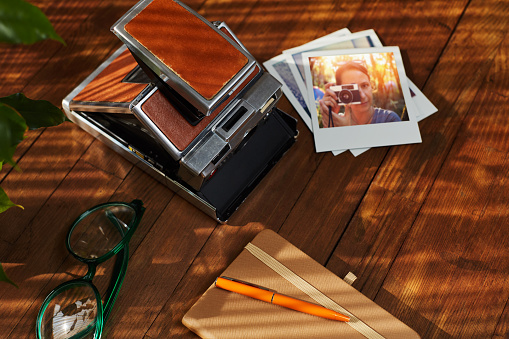Top view of a flat lay vintage instant film camera, printed photographs and notebook on wooden table under afternoon sunlight. Hipster items. The picture on instant photograph can be found in my portfolio Stock photo ID:862415084