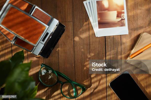Top View Of A Flat Lay Vintage Instant Film Camera Printed Photographs A Smart Phone And Notebook With Pen On Wooden Table Under Afternoon Sunlight With Copy Space In The Middle Hipster Items Stock Photo - Download Image Now