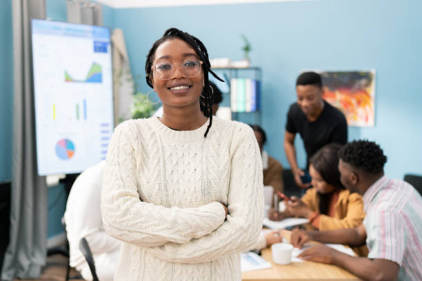 portrait of a smiling dark-skinned girl wearing a warm sweater with her arms crossed over chest, woman wears glasses and gold earrings, her hair is braided, a business meeting in office in background - hair net imagens e fotografias de stock