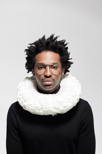 Afro american man wearing black turtleneck and white neck ruff, looking at camera. Headshot on grey background.