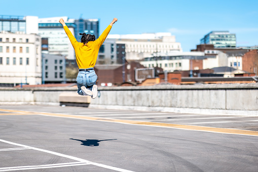 Successful black woman celebrating and jumping - Young woman wearing yellow top and curly hair celebrating victory and looking at city on background - happiness and success concepts
