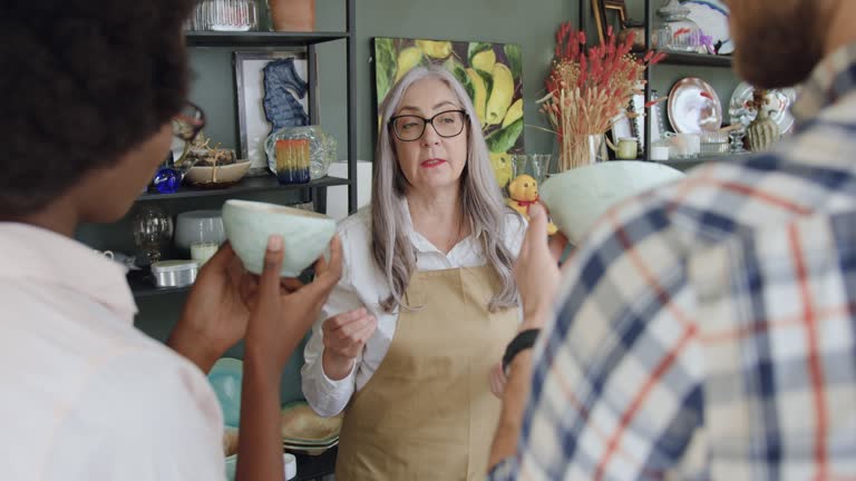 Good-looking friendly grey-haired mature seller proposing beautiful utensils to young mixed race couple which came in gift shop,front view