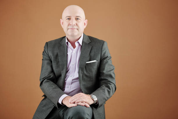 Portrait of content successful mature Caucasian man with bald head sitting with hands on knee against brown background