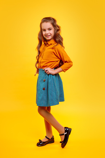 Side view full body preteen girl with long curly hair wearing trendy blouse and skirt standing, with hands on waist on yellow background and looking at camera with pretty smile