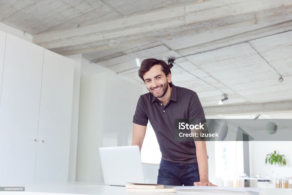Cheerful man in the office Happy mid adult men wearing dark polo shirt using computer in the modern white office. Man standing at the desk and smiling at camera. Polo Shirt Stock Photo