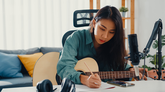 Happy asia woman songwriter play acoustic guitar listen song from smartphone think and write notes lyrics song in paper sit in living room at home studio.