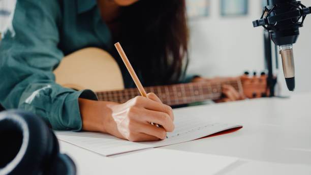 Happy asia woman songwriter play acoustic guitar listen song from smartphone think and write notes lyrics song in paper sit in living room at home studio. stock photo