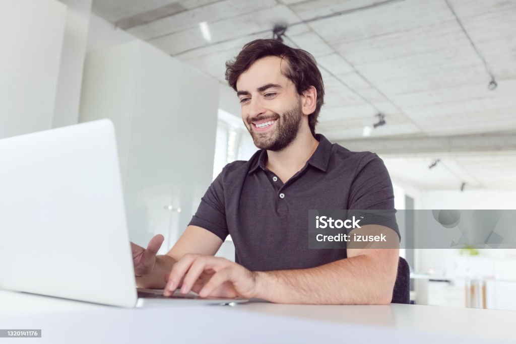 Cheerful man working in the office Happy mid adult men wearing dark polo shirt using computer in the modern white office. Man during video conference. Polo Shirt Stock Photo