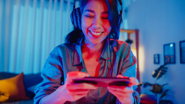 Happy asia girl gamer wear headphone competition play video game online with smartphone colorful neon lights in living room at home. Happy asia girl gamer wear headphone competition play video game online with smartphone colorful neon lights in living room at home. Esport streaming game online, Home quarantine activity concept. match lighting equipment photos stock pictures, royalty-free photos & images