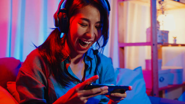 幸せなアジアの女の子のゲーマーは、自宅でカラフルなネオンライトのリビングルームでソファに座って友人とのスマートフォンの興奮した話でオンラインヘッドフォン競争ビデオゲームを� - portable player ストックフォトと画像