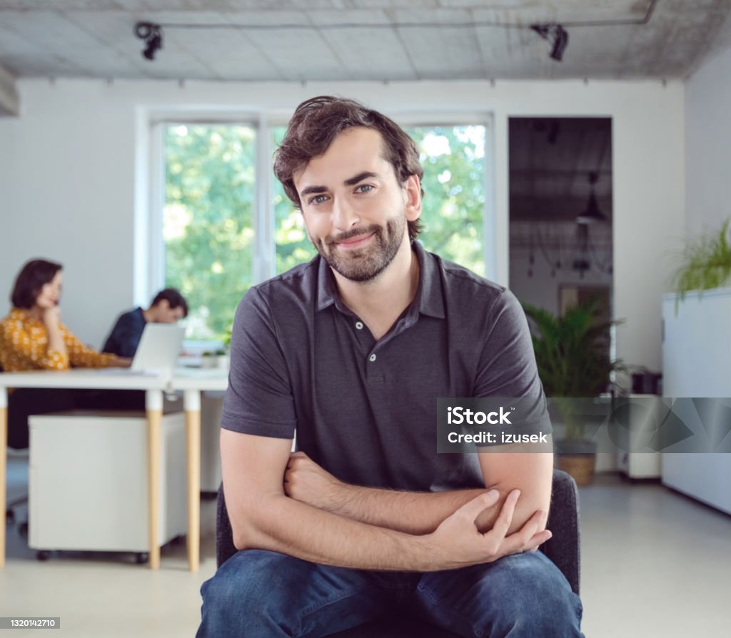 Portrait of man in the office Mid adult men wearing dark grey polo shirt sitting on chair in the modern white office, smiling at camera. Polo Shirt Stock Photo