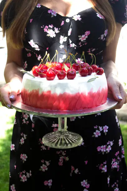 A beautiful young woman is holding and showing fruit cherry cake with white and red colors on a glass cake stand.