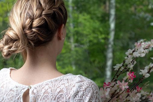A bride from behind, looking into the woods. Holding a bouquet of flowers.