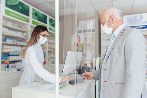 Shopping prescription drugs and pharmacist's advice, prescription drugs. A side view of a pharmacist standing behind a counter selling medicine to an mature man wearing a protective mask