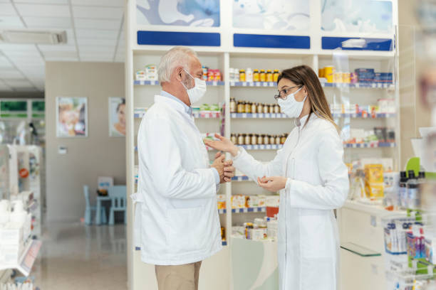 istock Side view of talking between two pharmacists and the corona virus. Before retiring, the pharmacist teaches the young pharmacist about working in a pharmacy. They wear uniforms and face masks 1320145784