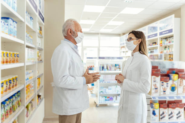 istock Talking between two pharmacists and the corona virus. Before retiring, the pharmacist teaches the young pharmacist about working in a pharmacy. They wear uniforms and face masks 1320145810
