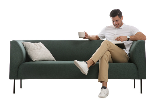 Man with cup of drink and book on comfortable green sofa against white background