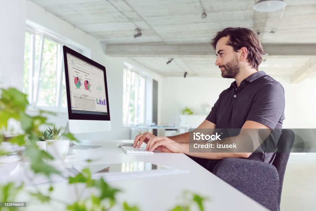 Man working in the office Thoughtful mid adult men wearing dark polo shirt using computer in the modern white office. Computer Monitor Stock Photo