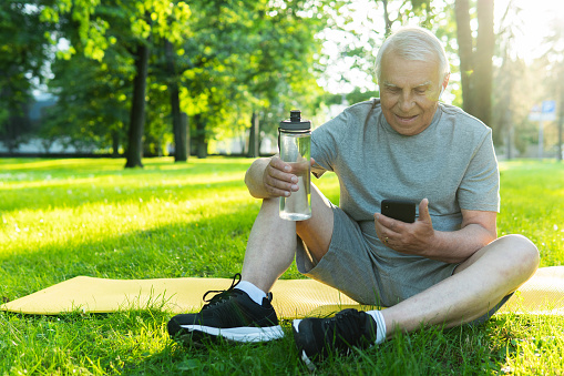 Calm elderly man with a smartphone and wireless earbuds relaxing after his workout in a city park