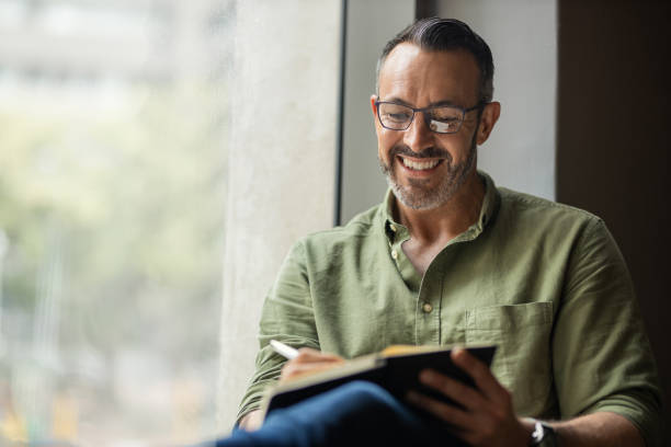 istock Mature man with beard, glasses writing in book sitting by sunny window 1320150014