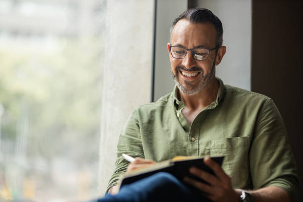 Mature man with beard, glasses writing in book sitting by sunny window Mature man with grey and black beard and glasses smiling, sitting near a bright sunny window writing in notebook with copy space diary stock pictures, royalty-free photos & images