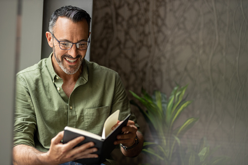 Mature man reading book by window, smiling wearing glasses, copy space