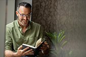 istock Mature man reading book by window, smiling wearing glasses, copy space 1320150020