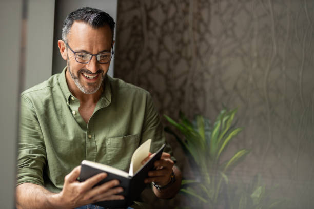 Mature man reading book by window, smiling wearing glasses, copy space Mature bearded man holding and reading book. Sitting next to sunny window, smiling and wearing glasses, copy space reading glasses stock pictures, royalty-free photos & images